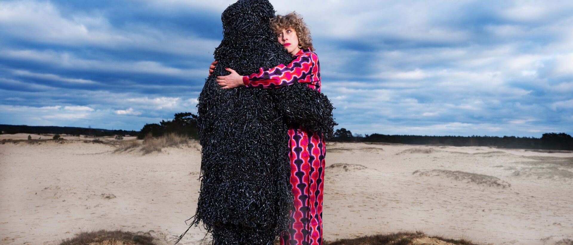 Twee acteurs staan op het strand en bij een van hun is de kleding gemaakt van het lint van cassettebandjes. Ze omhelzen elkaar