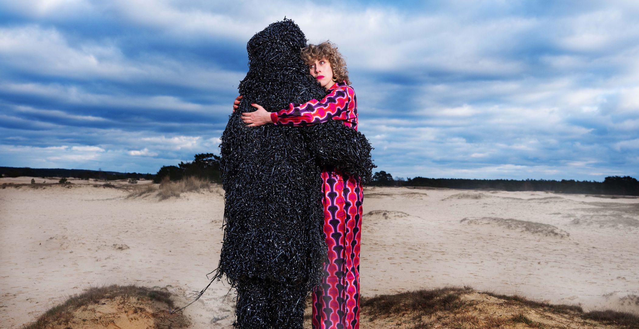 Twee acteurs staan op het strand en bij een van hun is de kleding gemaakt van het lint van cassettebandjes. Ze omhelzen elkaar