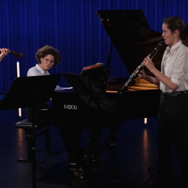 drie personen zijn verschillende instrumenten aan het bespelen. Eentje speelt piano, nog een speelt viool en de derde speelt ene fluitinstrument