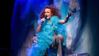 Milène van der Smissen heeft een extravagante blauwe jurk aan, ze zingt in een microfoon met haar haren wapperend in de wind. Het is duidelijk op het toneel.