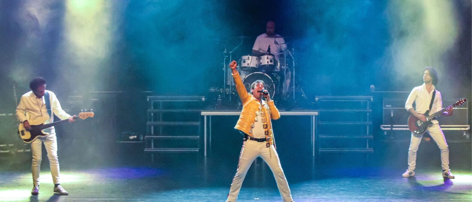 Er staan 4 Mannen op de foto 1 is aan het drummen, er zijn 2 mannen aan het gitaar spelen en in het midden is iemand aan het zingen. Er zijn blauwe spotlights