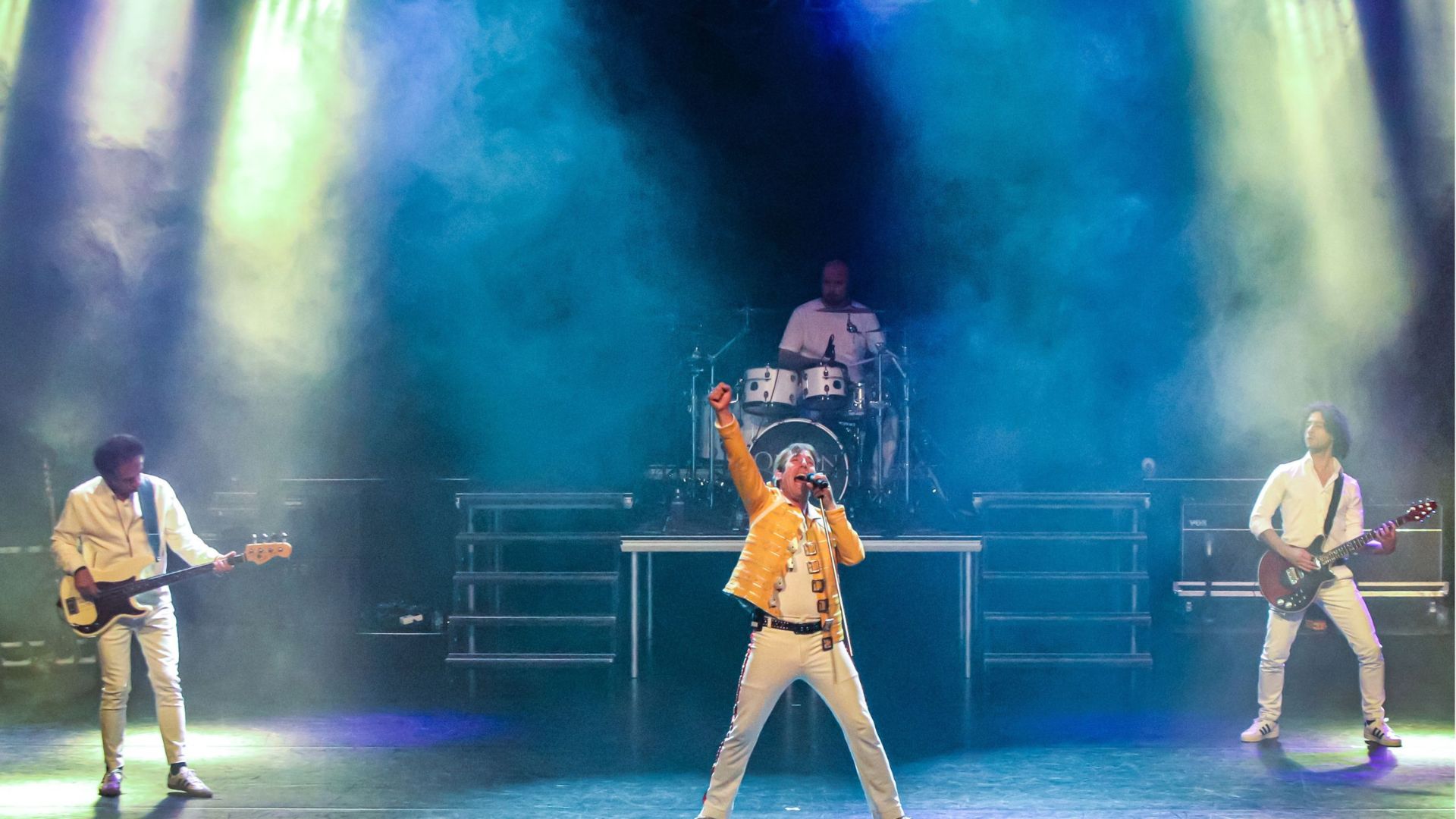 Er staan 4 Mannen op de foto 1 is aan het drummen, er zijn 2 mannen aan het gitaar spelen en in het midden is iemand aan het zingen. Er zijn blauwe spotlights