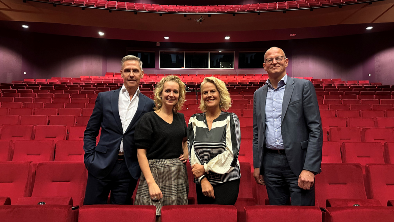 Vier mensen kijken lachend de camera in, ze staan in een theaterzaal tussen de rode stoelen.