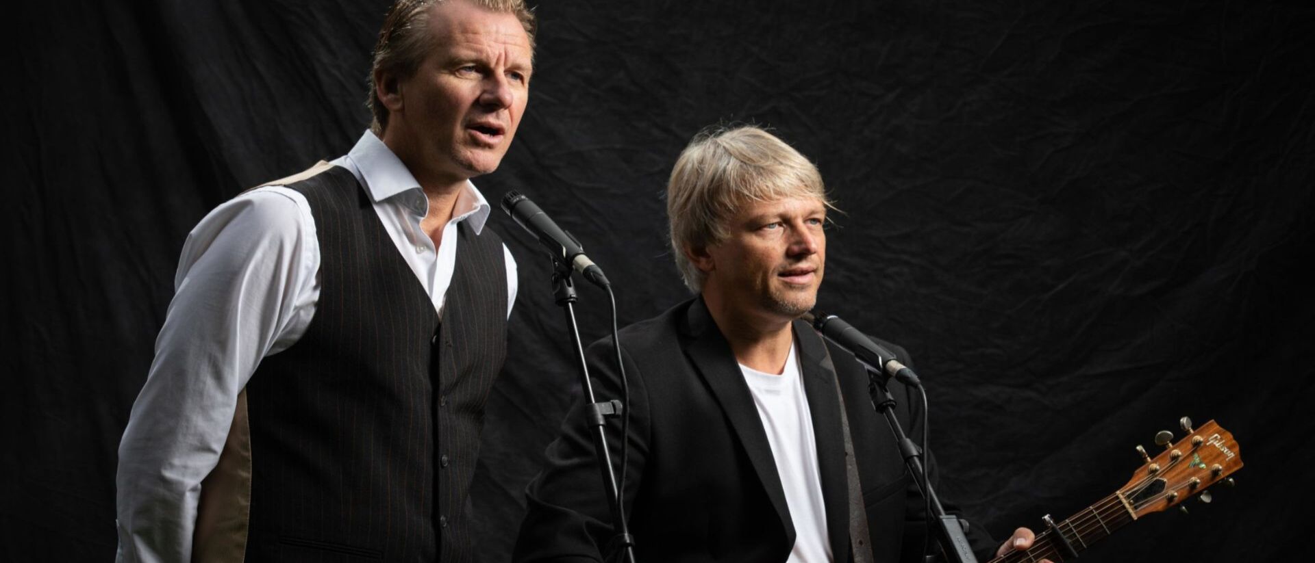 Niels van der Gulik en Frank Kooijman staan voor zwarte achtergrond met gitaar en microfoon.