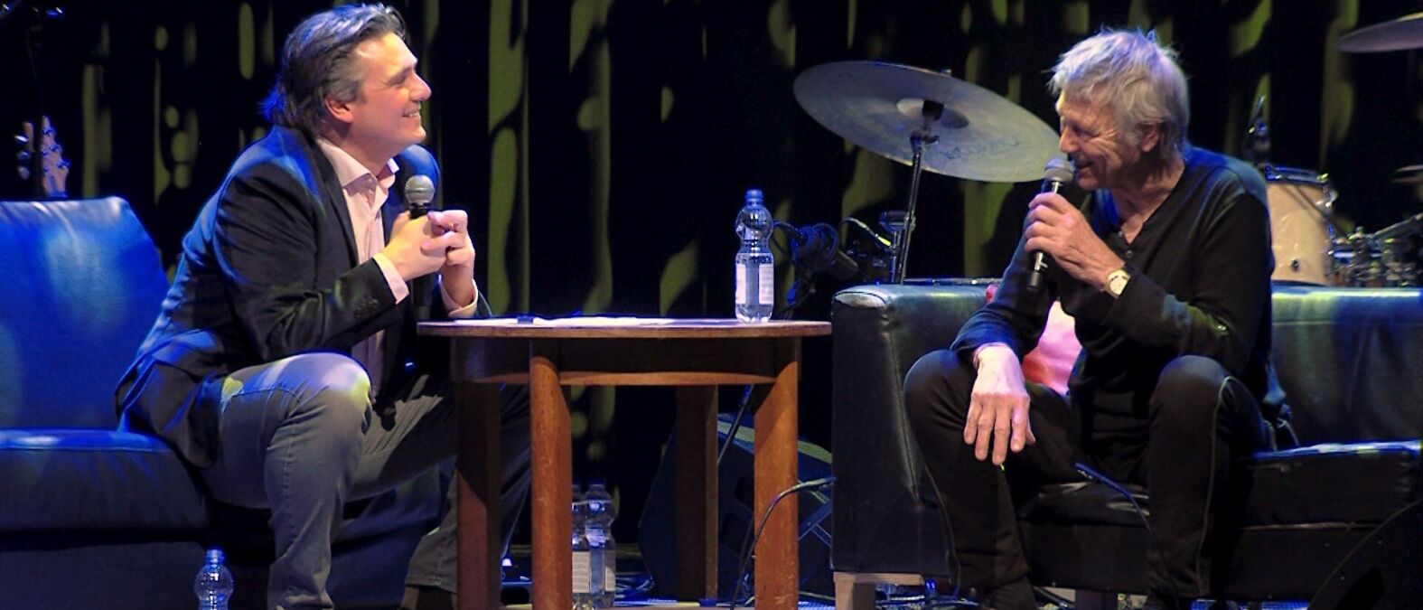 Twee mannen zitten op een podium met allebei een microfoon in hun hand