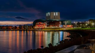 Foto in de avond genomen van het gebouw van Schouwburg Het Park met het water van het Markermeer ervoor. Door de schemering en alle lichten die aan staan, ziet het gebouw en prachtig uit