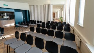 Vergaderzaal met rijen zwarte stoelen en een opening naar een andere zaal