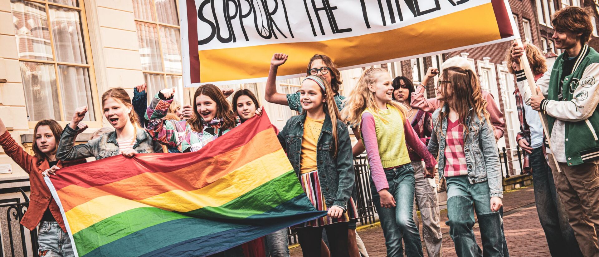 Kinderen protesteren vrolijk. Ze hebben een regenboogvlag vast en een protestbord waar 'Support the miners' op staat.