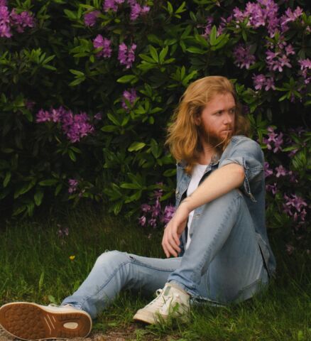 Man met rood lang haar zittend op gras met paarse bloemen achtergrond.