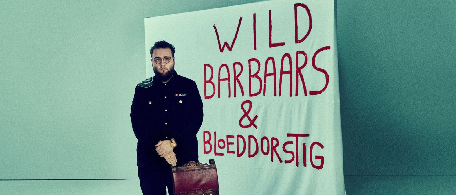 Sezgin Gulec staat met een stoel voor zich en op de achtergrond een wit doet en met rode letters: Wild Barbaars & Bloeddorstig