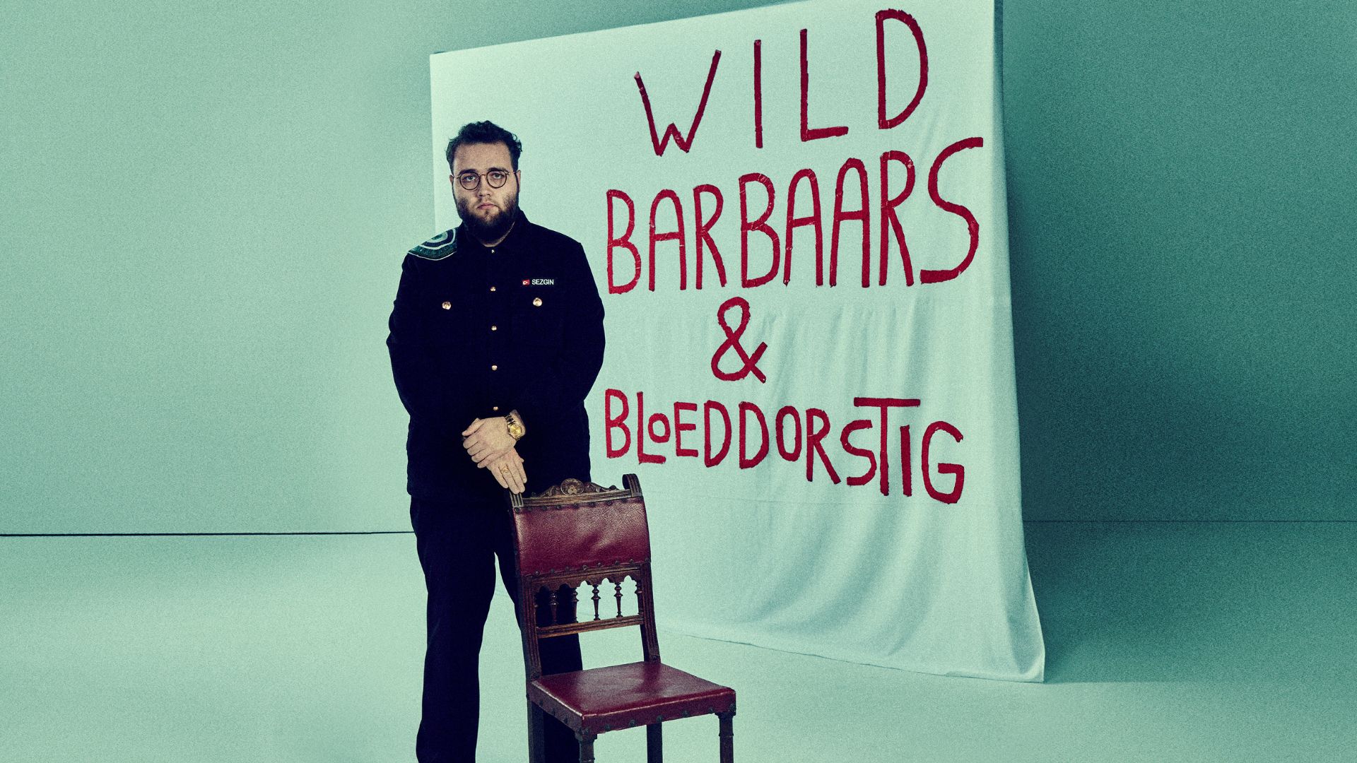 Sezgin Gulec staat met een stoel voor zich en op de achtergrond een wit doet en met rode letters: Wild Barbaars & Bloeddorstig