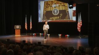 Een foto van Raymond Mens op een podium met Amerikaanse vlaggen en hij projecteert een beeld.