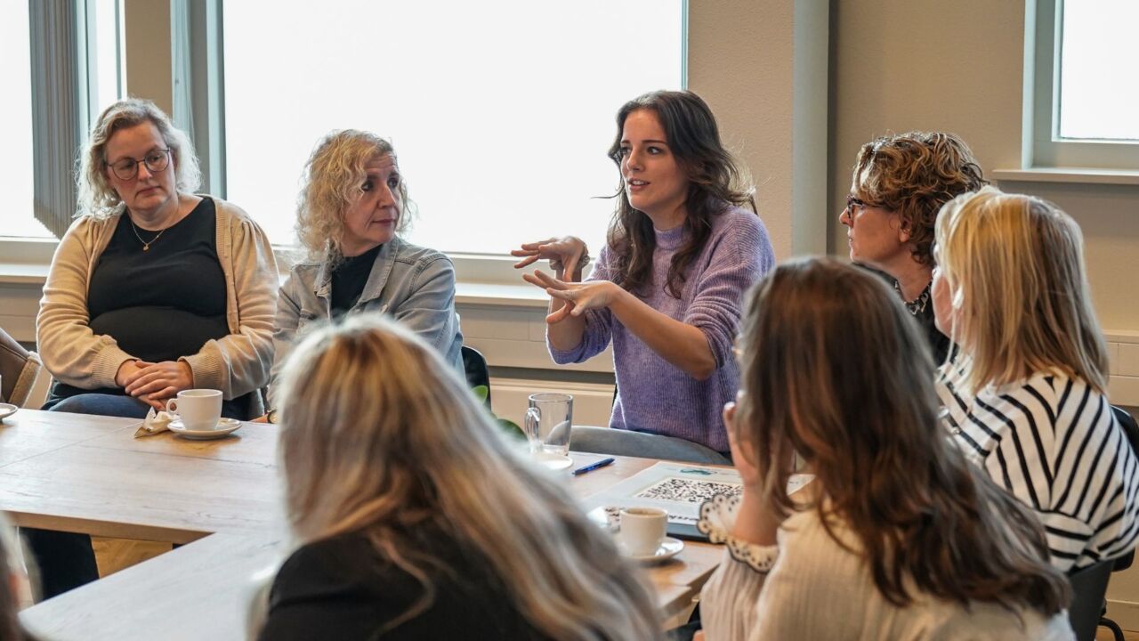 Een groep vrouwen zit in een halve cirkel om een tafel heen en vergadert