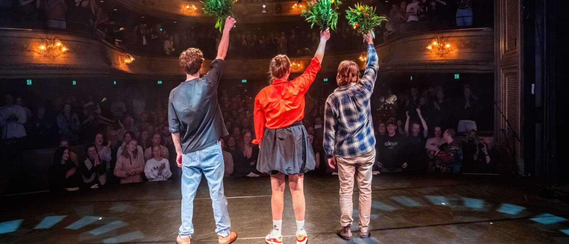 twee mannen en een vrouw staan op het podium op het punt een buiging te doen. De foto is van de achterkant genomen waardoor je de volle zaal ziet.