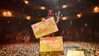 Foto van de grote winnaar van het Leids Cabaret festival. Met zijn rug naar het publiek met een grote glimlach op zijn gezicht en beker in zijn handen.