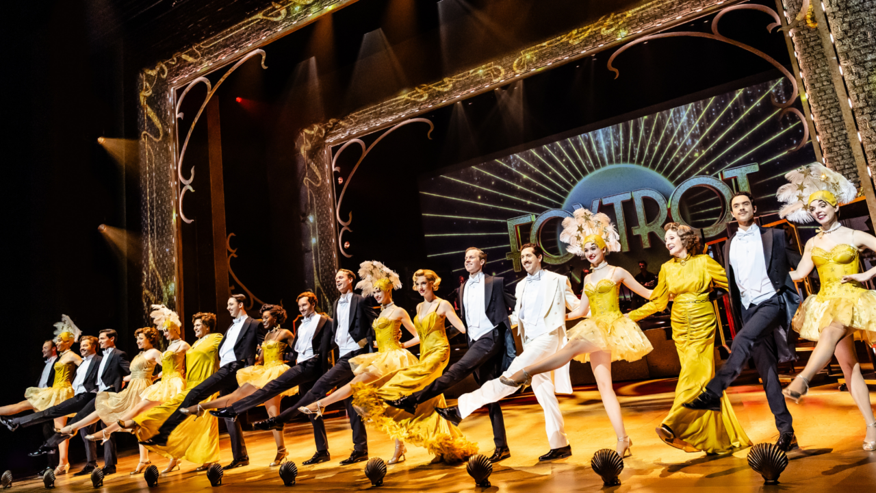 De cast hand in hand naast elkaar te dansen, de mannen in pak en de vrouwen in een mooie gouden jurk