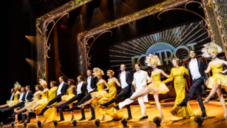 De cast hand in hand naast elkaar te dansen, de mannen in pak en de vrouwen in een mooie gouden jurk