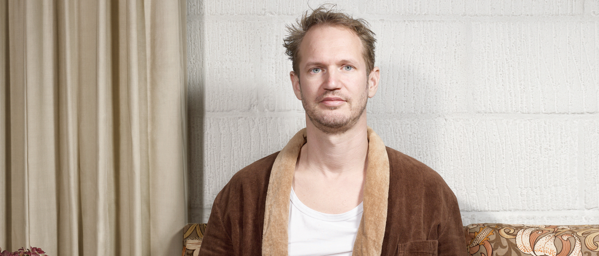 Man met warrig haar zit op een retrobank, draagt een bruine badjas en wit hemd, en kijkt recht in de camera in een huiselijke setting met neutrale achtergrond.