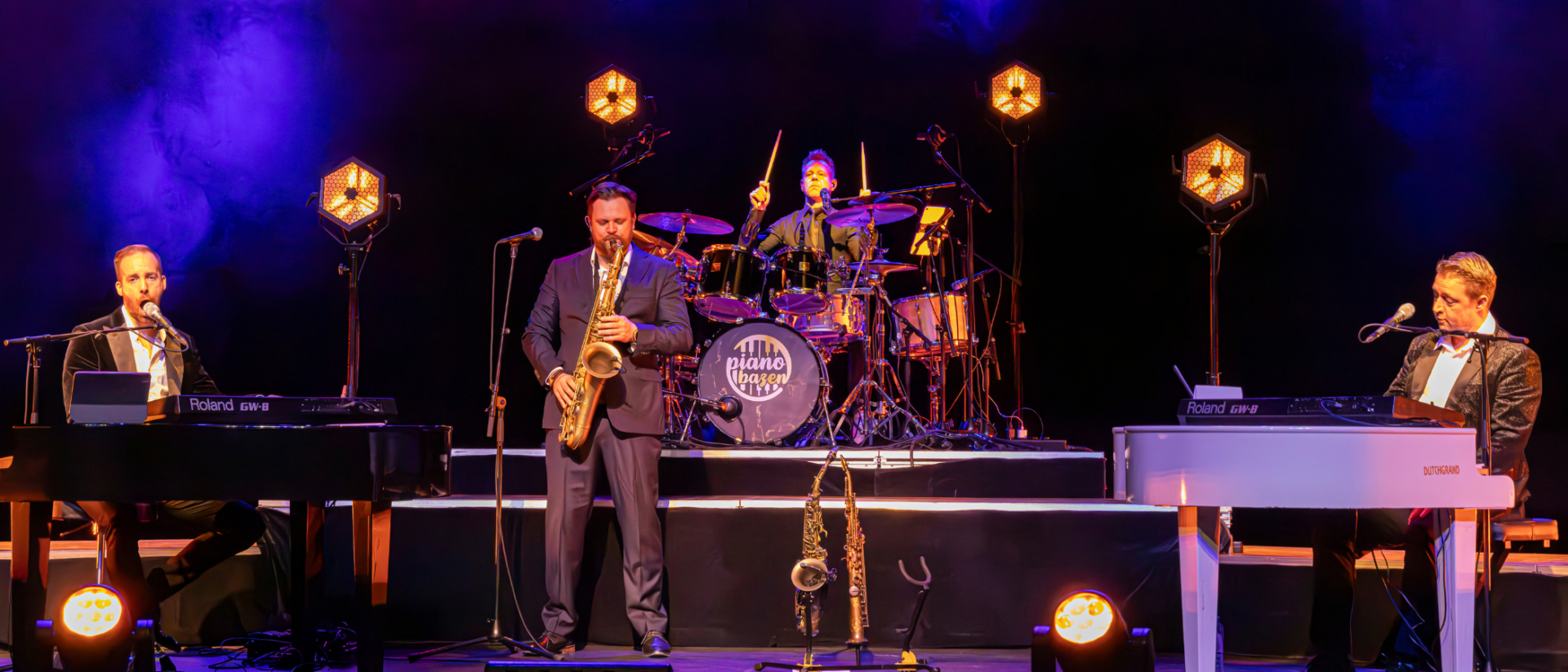 De Pianobazen live op het podium, met twee pianisten, een saxofonist en een drummer in kleurrijk licht tijdens een theatervoorstelling.