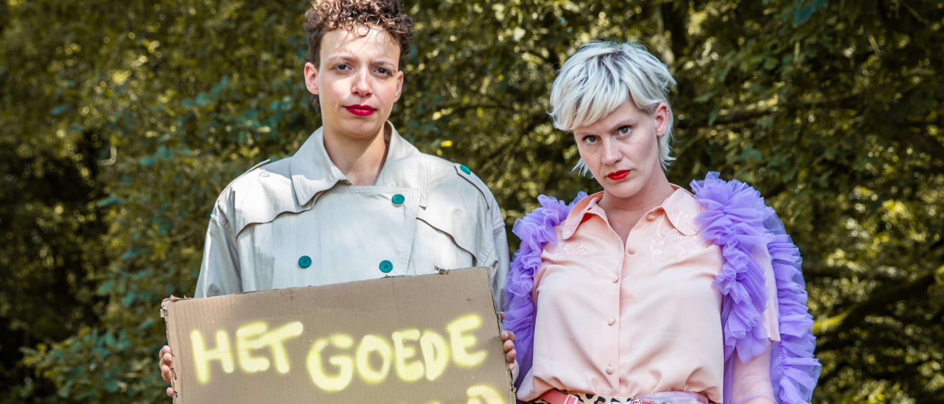 Twee vrouwen poseren in kleurrijke en contrasterende outfits in een bosrijke omgeving. Eén van hen houdt een kartonnen bord vast met de tekst 'HET GOEDE VOORBEELD' in gele graffitiletters.