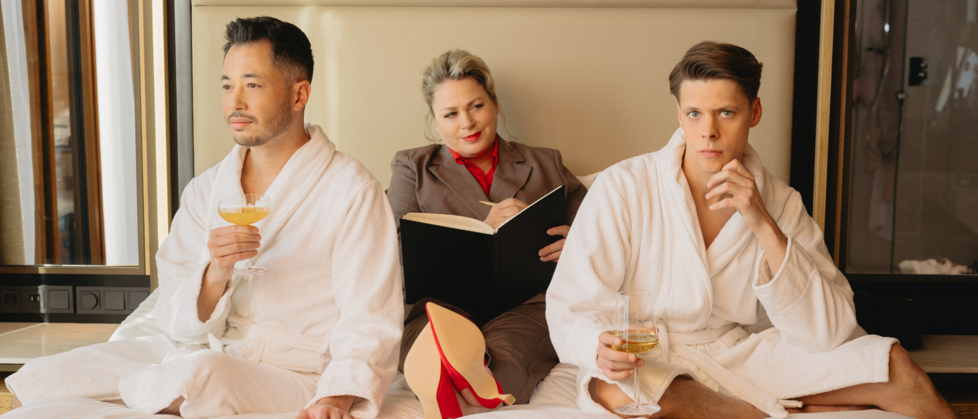 Twee mannen in witte badjassen zitten op een bed met een drankje, terwijl een vrouw in pak tussen hen in zit en aantekeningen maakt in een boek.