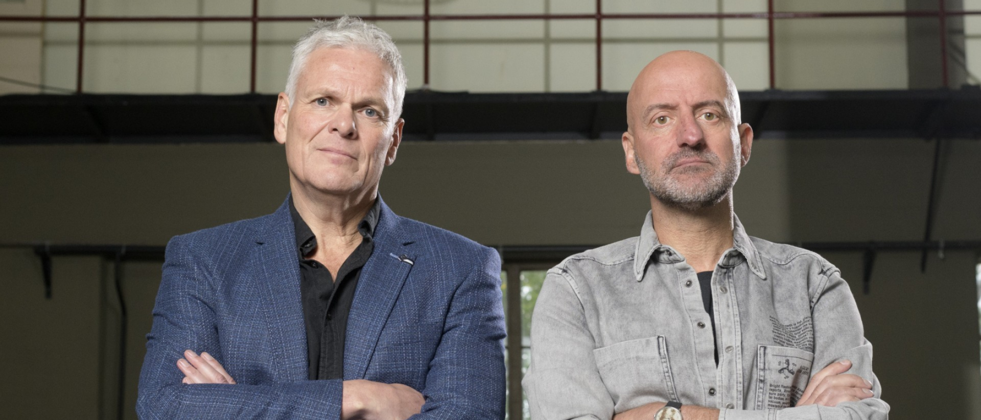 Twee mannen zitten met armen over elkaar op stoelen in een industriële ruimte, kijkend naar de camera met een serieuze blik.
