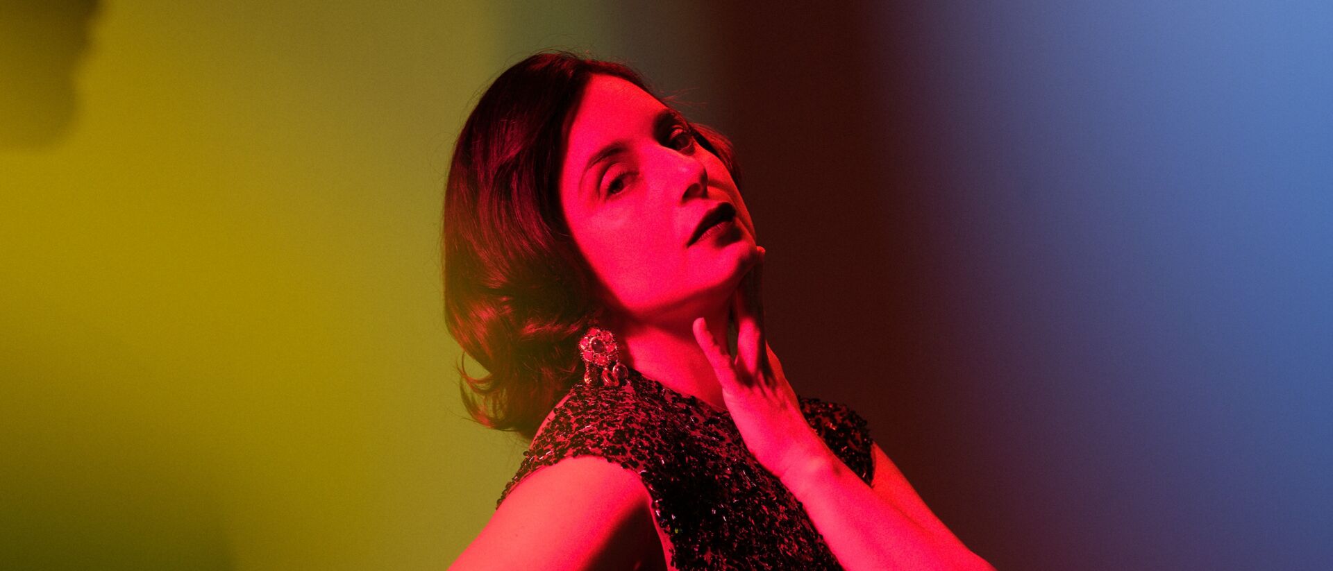 Portret van een vrouw in artistiek gekleurd licht met rode, gele en blauwe tinten, gekleed in een glinsterende jurk en kijkend in de camera.