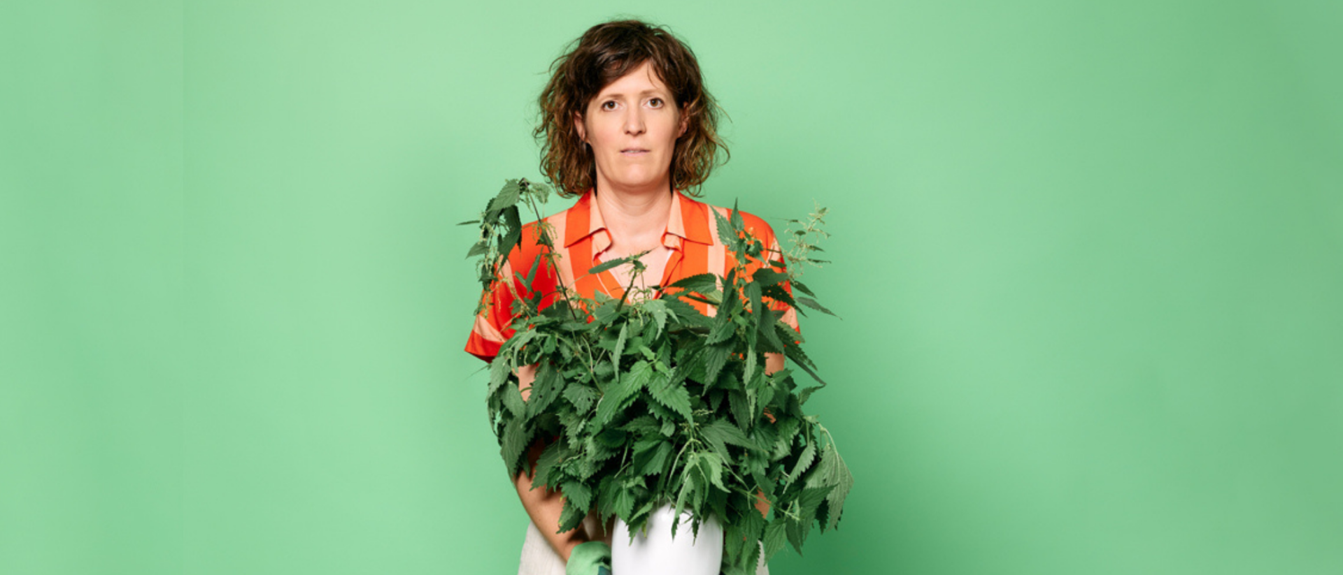 Janneke de Bijl tegen een lichtgroene achtergrond. Ze kijkt onzeker met een wietplant in haar handen