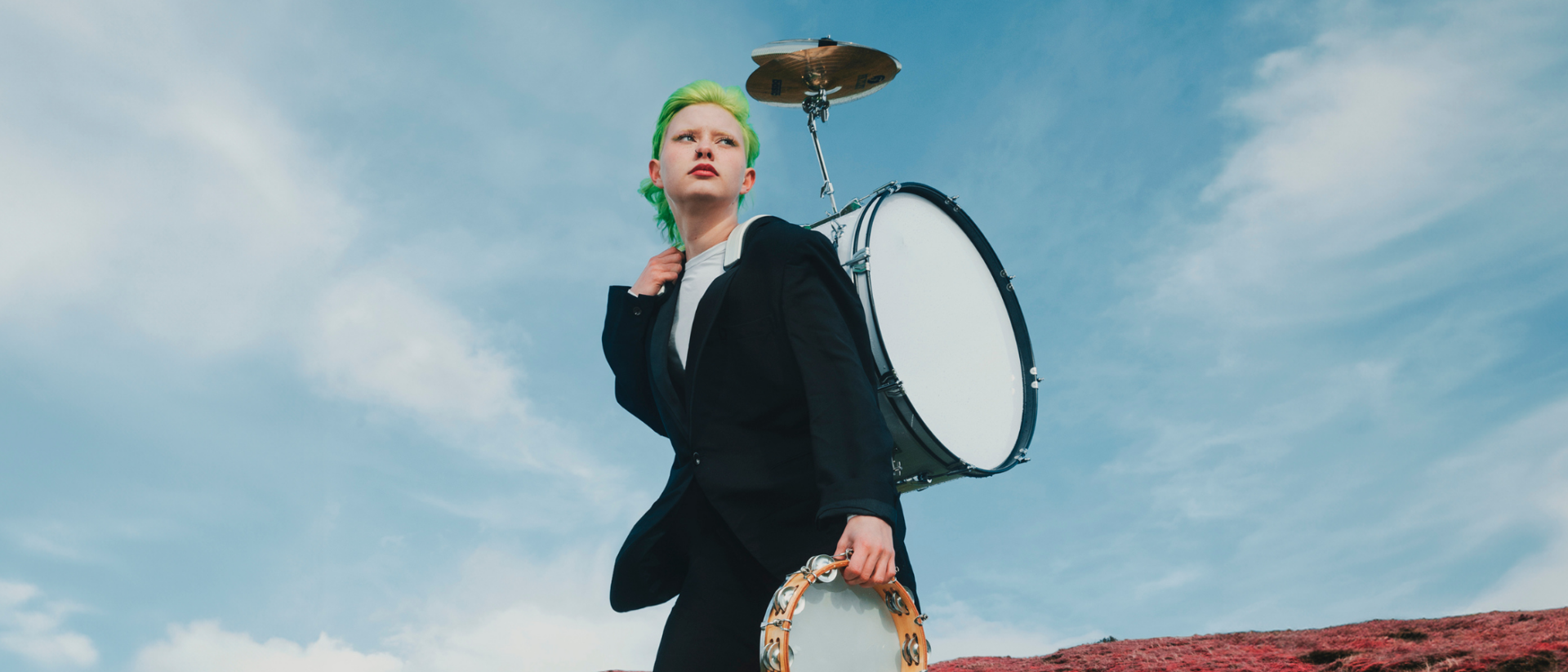 Jonge performer met groen haar, drumstel op de rug en tamboerijn in de hand, staand op een rood veld onder een blauwe lucht.