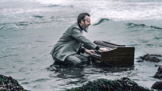 Man in pak speelt piano in zee bij rotsen – De Wilde Noordzee Hoorn