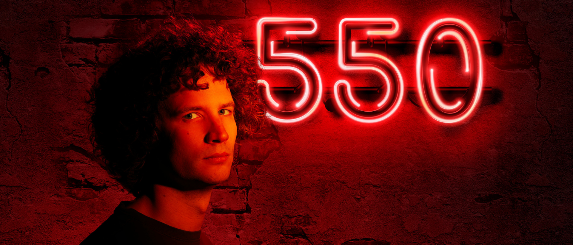 Man met krullend haar in rood licht kijkt indringend naar de camera. Naast hem staat het getal 550 in felrode neonletters op een verweerde bakstenen muur.