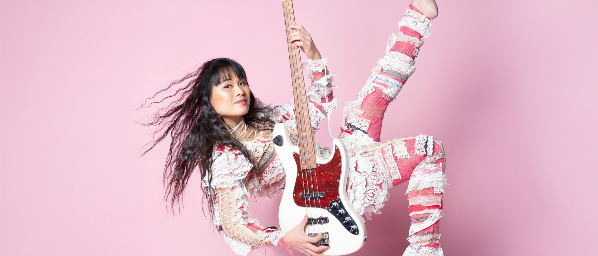 Vrouw in kleurrijk patchworkkostuum zweeft speels in de lucht met een witte basgitaar, voor een roze achtergrond.