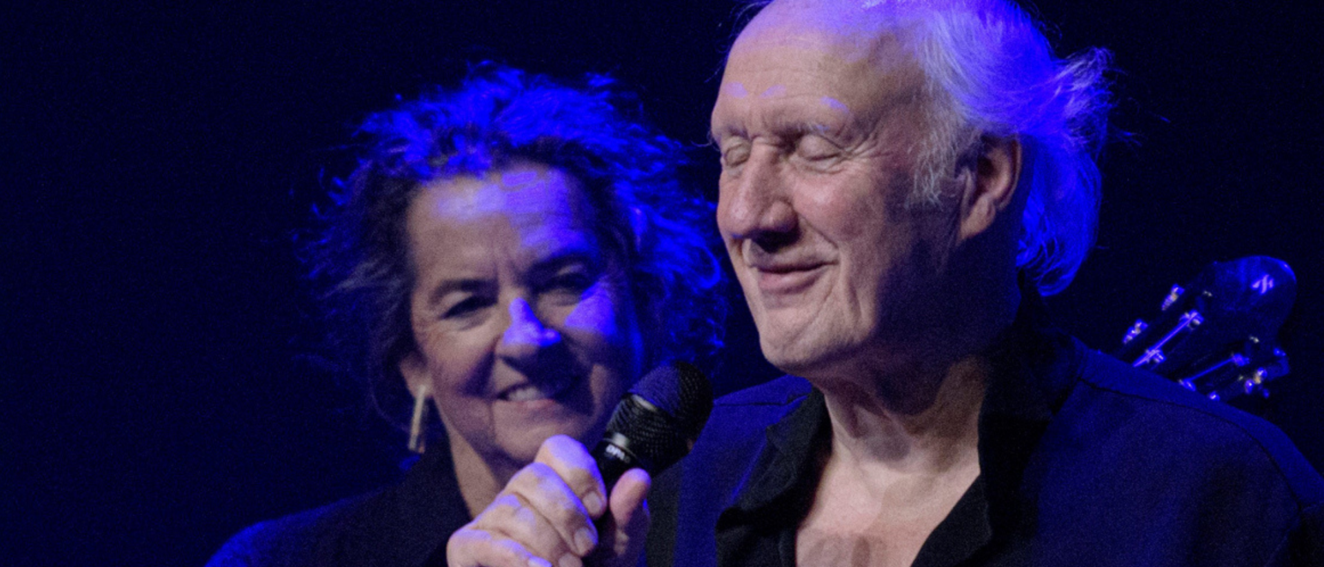 Oudere man zingt met gesloten ogen in microfoon, terwijl een vrouw met grijze krullen en warme glimlach op de achtergrond toekijkt, beiden verlicht door blauw podiumlicht.