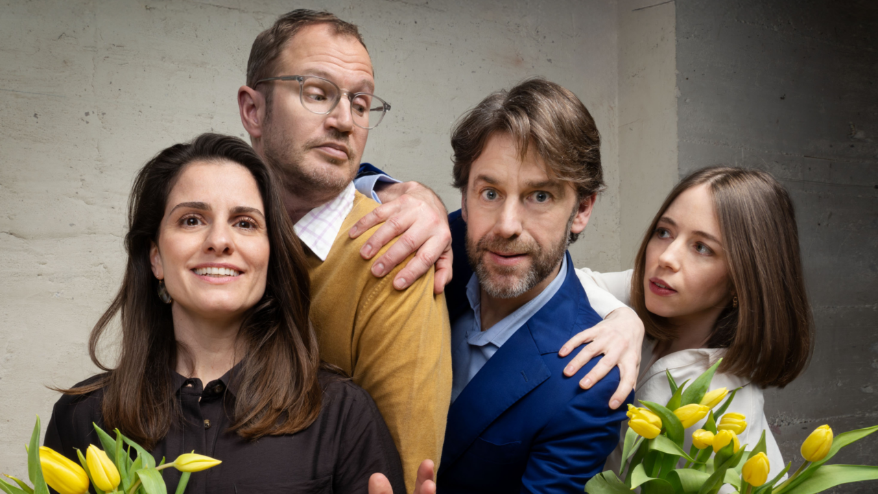 Vier volwassenen poseren in een licht komische setting met gele tulpen in hun handen; twee mannen en twee vrouwen staan dicht bij elkaar voor een betonnen muur.