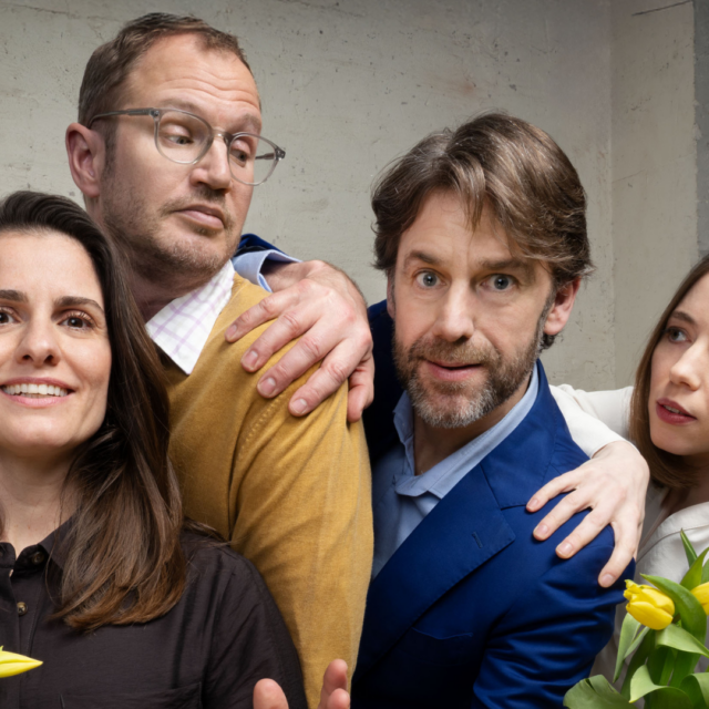 Vier volwassenen poseren in een licht komische setting met gele tulpen in hun handen; twee mannen en twee vrouwen staan dicht bij elkaar voor een betonnen muur.