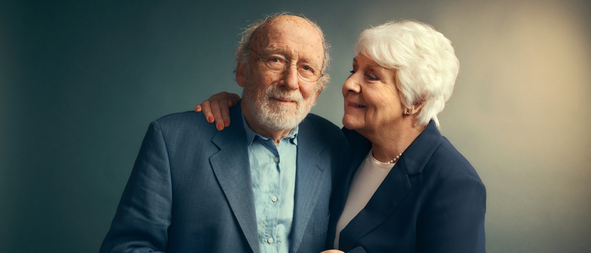 Een man en een vrouw staan lief tegen elkaar aan te poseren voor de foto. De vrouw kijkt de man aan terwijl de man naar de camera kijkt.