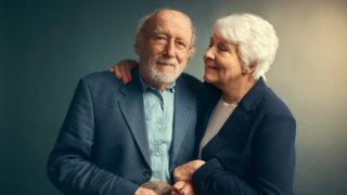 Een man en een vrouw staan lief tegen elkaar aan te poseren voor de foto. De vrouw kijkt de man aan terwijl de man naar de camera kijkt.