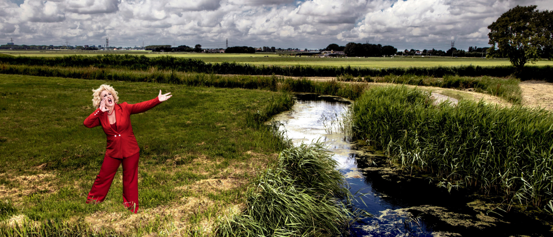 Een vrouw in een felrood pak staat schreeuwend in een uitgestrekt Hollands polderlandschap met sloot, weiland en wolkenlucht. Ze houdt één hand aan haar mond en de andere gestrekt in de lucht, alsof ze een boodschap roept over het land.