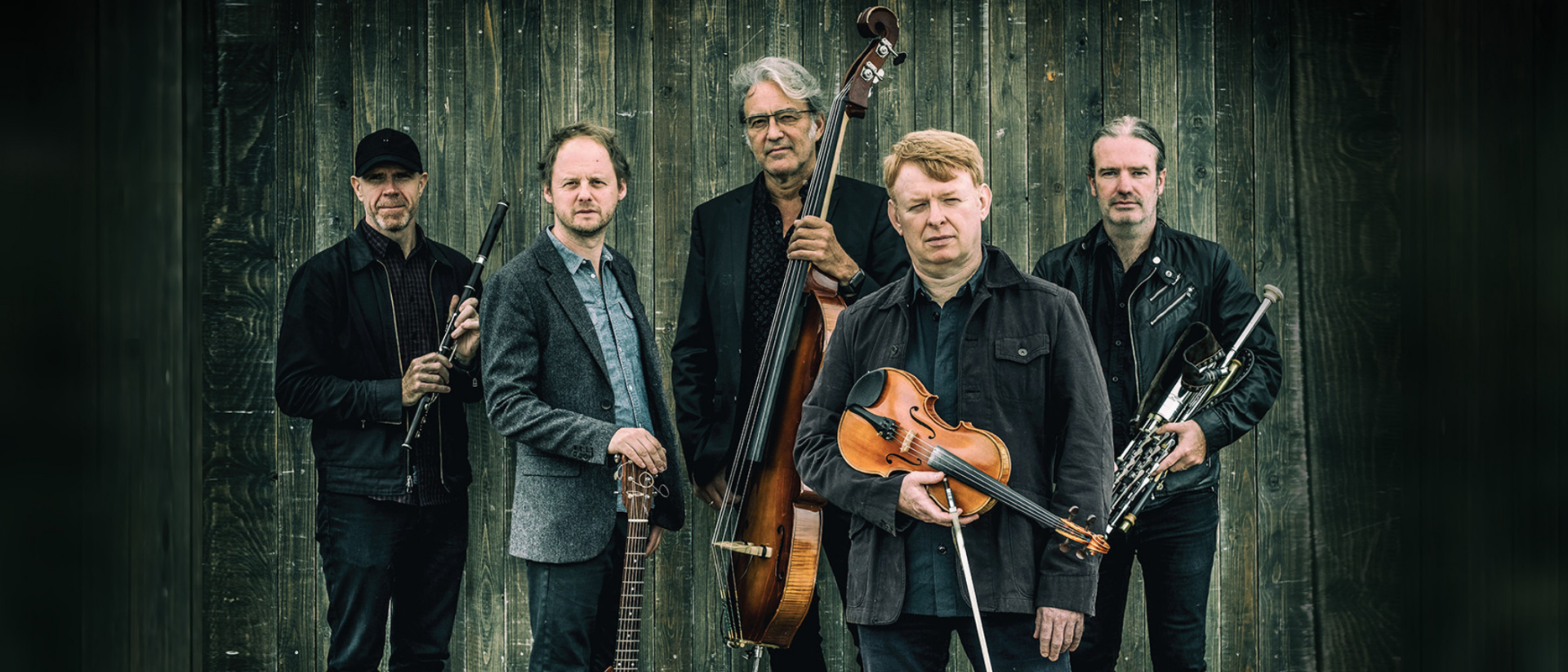 Vijf mannen poseren met hun muziekinstrumenten — viool, gitaar, contrabas, fluit en doedelzak — voor een houten achtergrond, gekleed in donkere kleding.