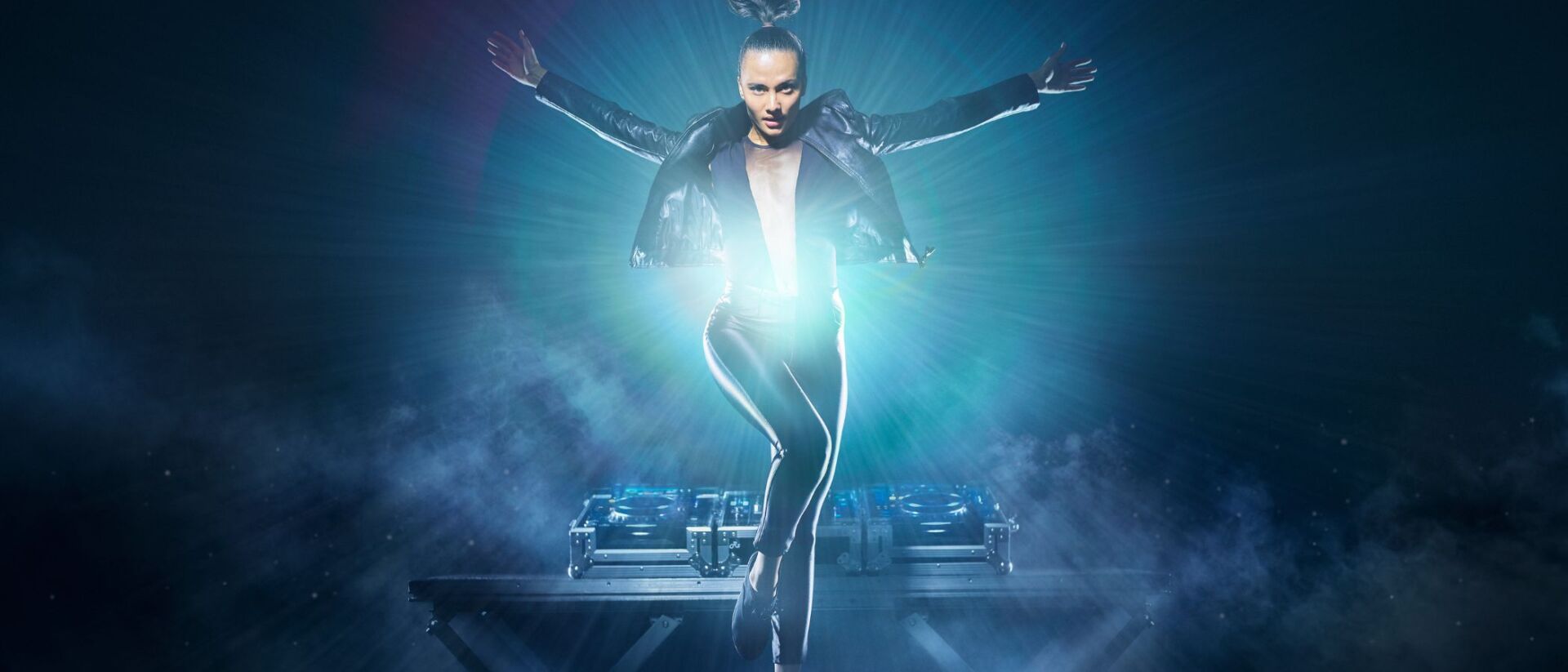 Dansperformer in leren jas springt boven een dj-tafel, verlicht door een zachte lichtstraal.