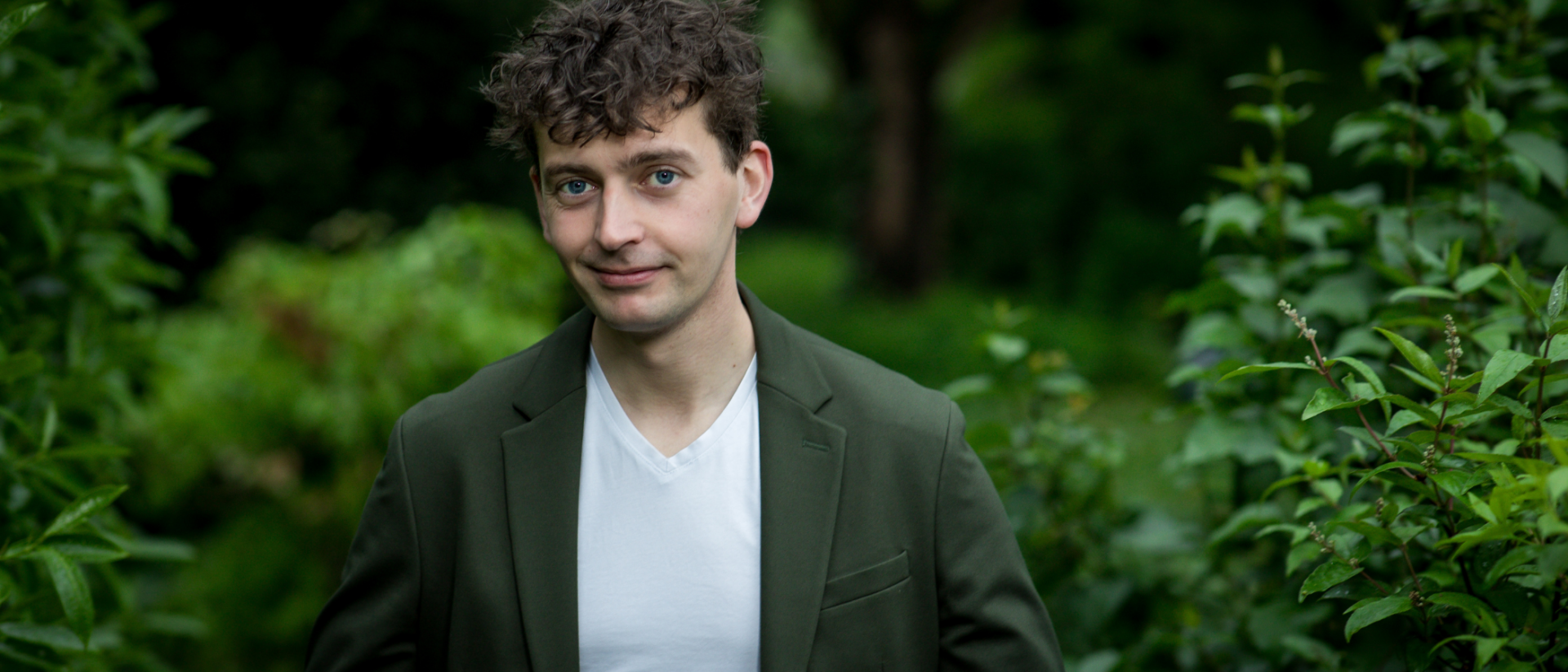 Man met krullend haar en groene blazer staat in een weelderige, groene tuin en kijkt met een lichte glimlach in de camera.