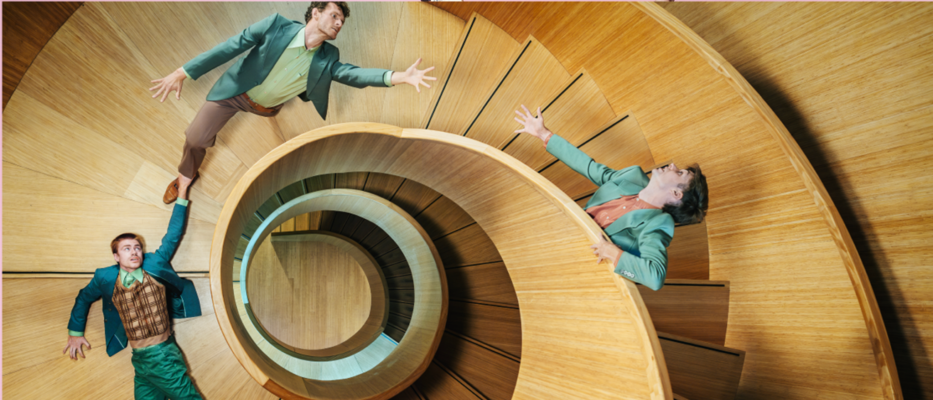 Drie mannen in kleurrijke pakken poseren op verschillende niveaus van een spiraalvormige houten trap, wat een surrealistisch en speels perspectief oplevert.