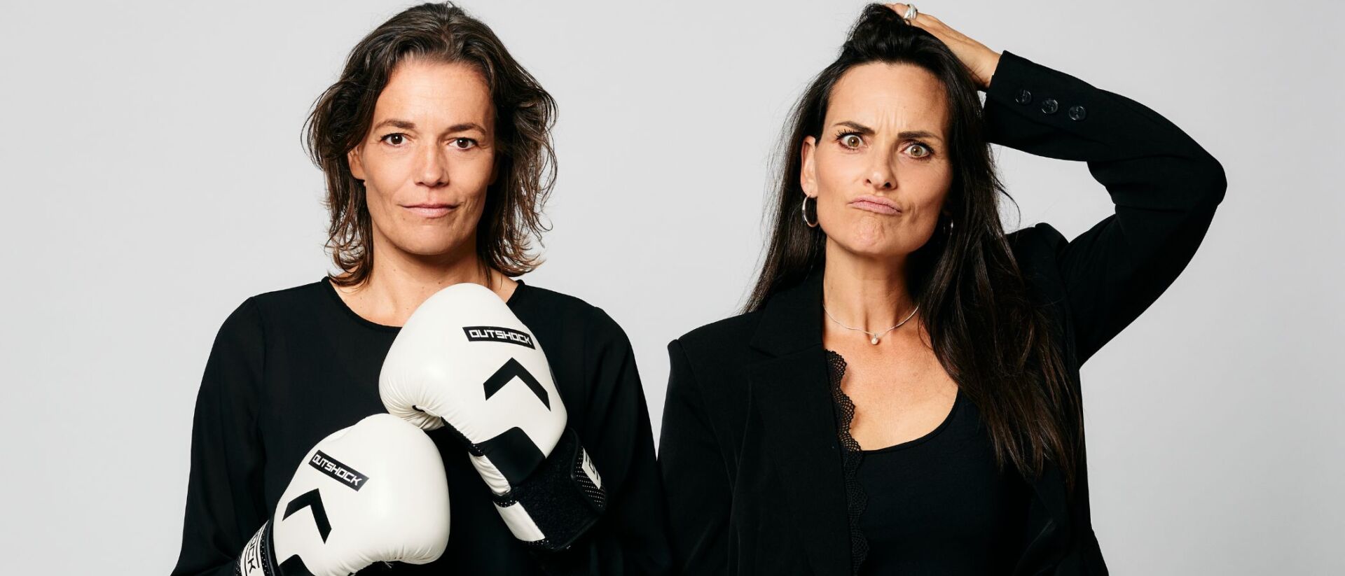 je ziet Marieke van Ginneken en Ilse van den Heuvel naast elkaar op de afbeelding staan. De een heeft bokshandschoenen aan en de ander gaat met een hand door haar haren.