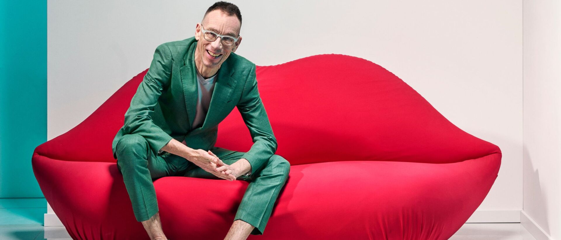 Dolf Jansen in een groen pak zit ontspannen op een felrode lipvormige bank, met een glimlach, op een moderne, minimalistische achtergrond.