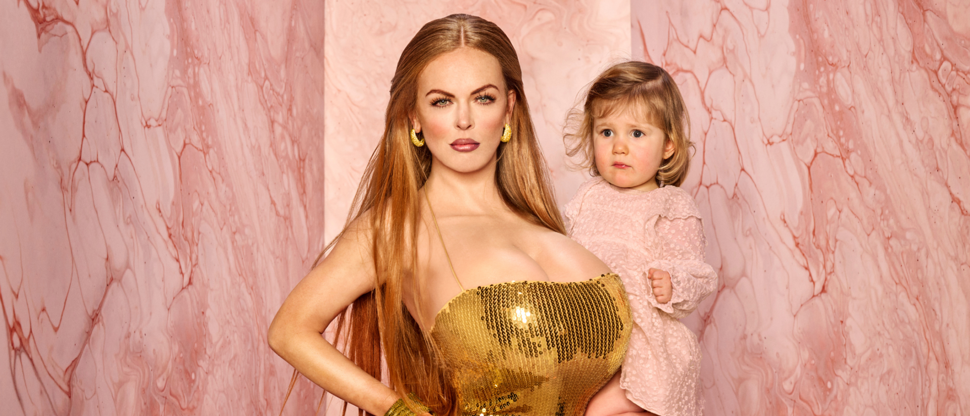Vrouw in een goudkleurige glitterjurk poseert krachtig met een jong kind op de arm, tegen een roze marmerachtige achtergrond.