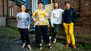 Groepsfoto van vier muzikanten in kleurrijke kleding, poserend voor een bakstenen muur, met een ontspannen en vrolijke uitstraling