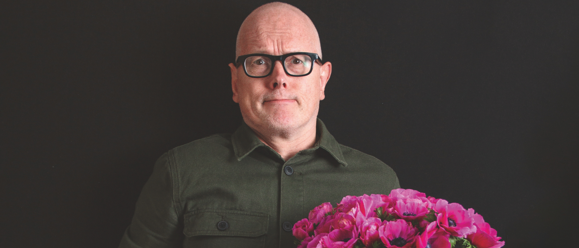 Kale man met bril staat voor een zwarte achtergrond, houdt een bos felroze bloemen in de ene hand en een roze retro wekker in de andere, met een licht verbaasde blik.