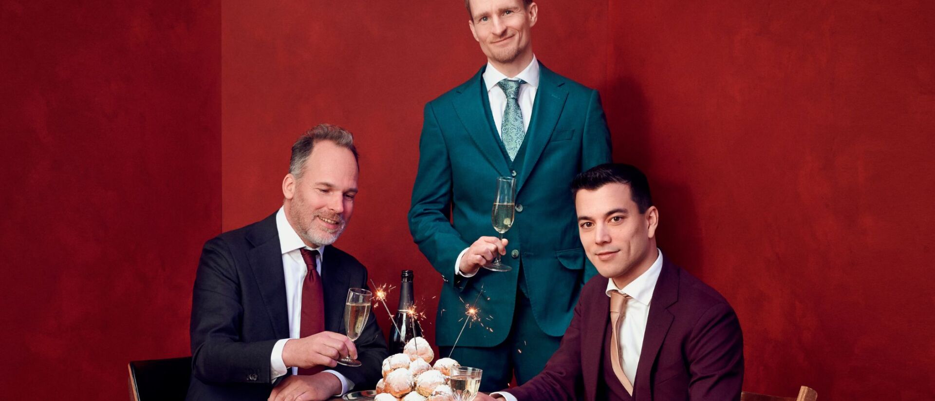 3 mannen in stijlvolle pakken proosten met champagne en houden een schaal met oliebollen vast, terwijl er vuurwerk aanstekers uit de oliebollen komen. De achtergrond is donkerrood.