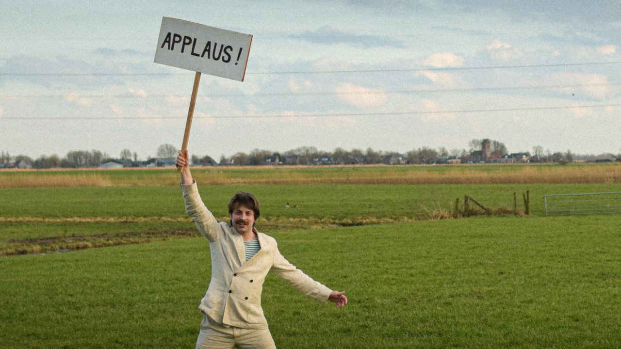 Man in licht pak staat juichend in een uitgestrekt weiland en houdt een bord omhoog met de tekst “APPLAUS!” tegen een achtergrond van Hollandse wolkenlucht en hoogspanningsmast.