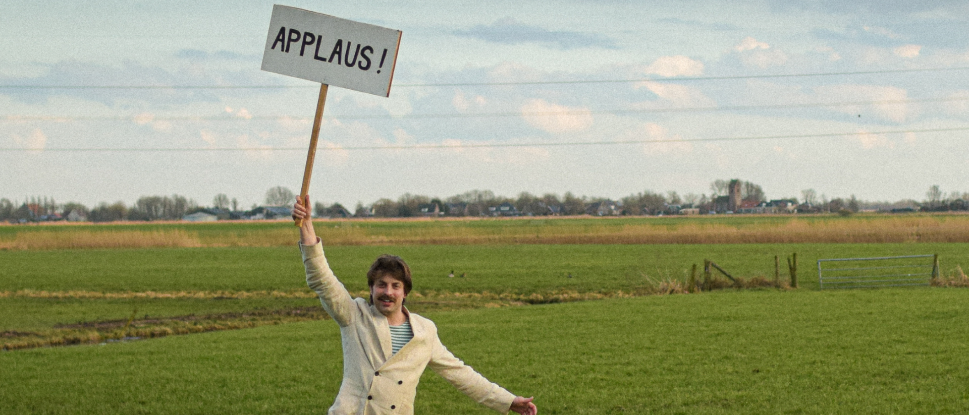 Man in licht pak staat juichend in een uitgestrekt weiland en houdt een bord omhoog met de tekst “APPLAUS!” tegen een achtergrond van Hollandse wolkenlucht en hoogspanningsmast.